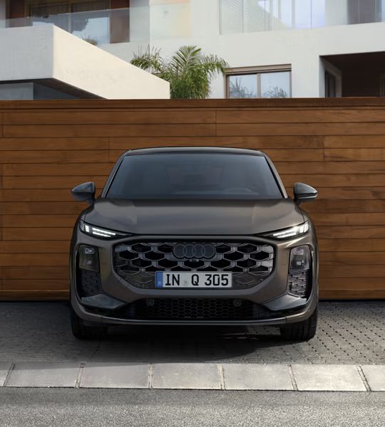 Der Q3 Sportback in Frontansicht parkt vor einem modernen Gebäude, dahinter eine Wand in Holzoptik.