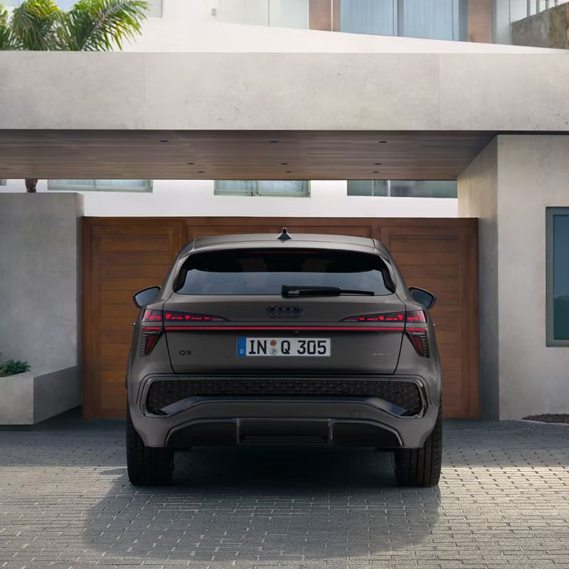 Der Audi Q3 Sportback ist in einer Heckansicht zu erkennen. Er steht auf einem Parkplatz vor einem hölzernen Eingangstor zu einem Wohnhaus.