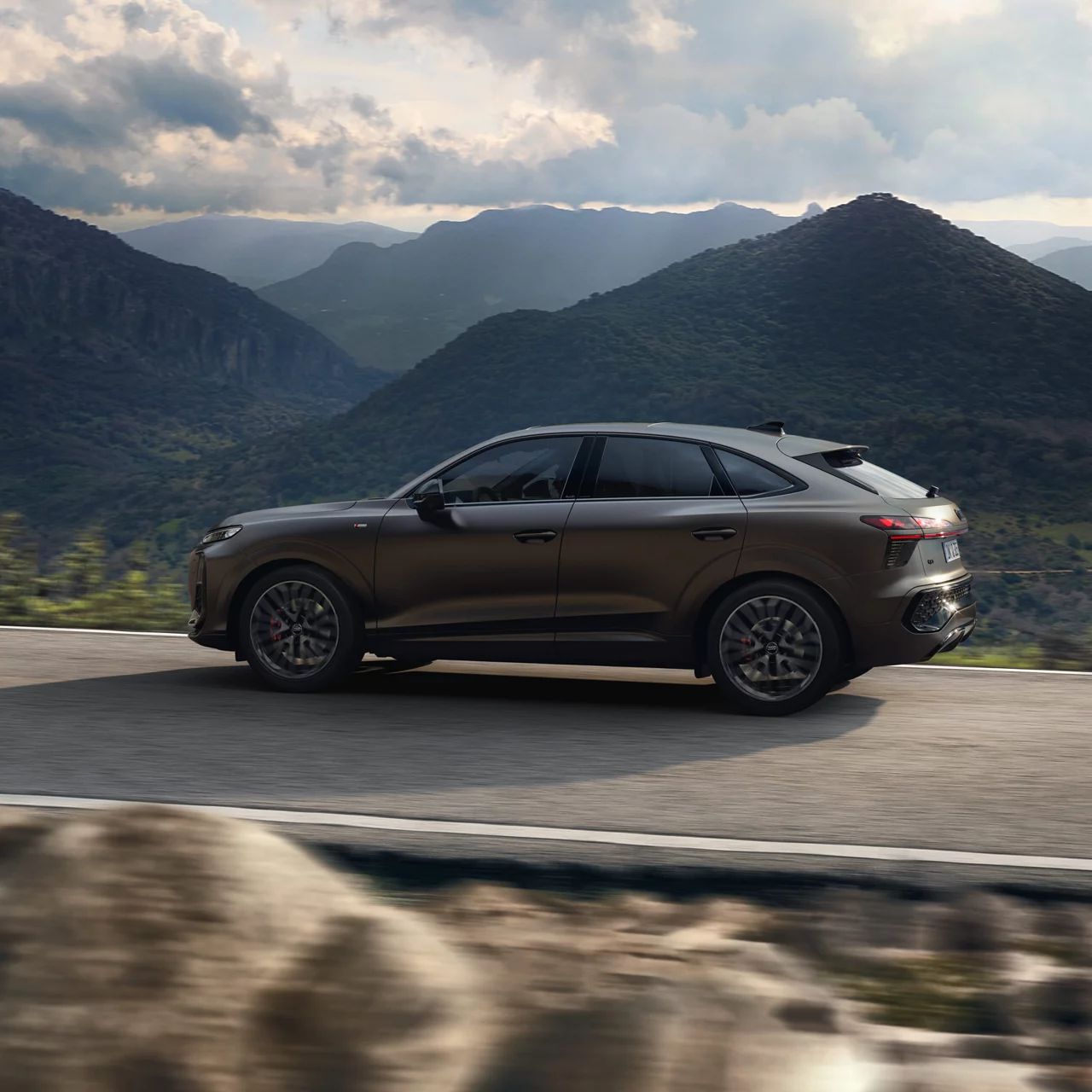 Ein brauner Audi Q3 Sportback fährt auf einer kurvigen Straße durch eine bergige Landschaft. Im Hintergrund sind bewaldete Hügel und ein bewölkter Himmel zu sehen. 