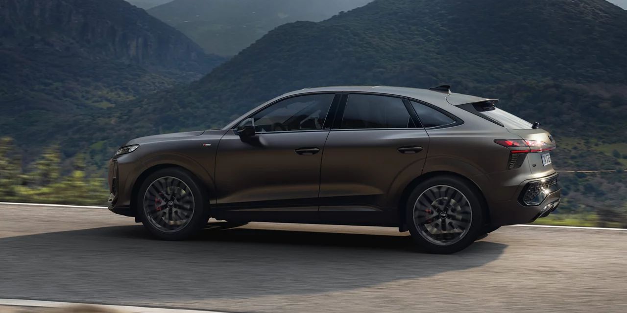 Ein brauner Audi Q3 Sportback fährt auf einer kurvigen Straße durch eine bergige Landschaft. Im Hintergrund sind bewaldete Hügel und ein bewölkter Himmel zu sehen. 